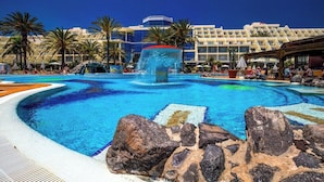 A large outdoor swimming pool with a central water feature, surrounded by palm trees and a hotel building in the background. - Sbh Costa Calma Palace (Pajara)