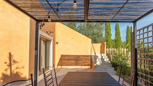 Un espace extérieur abrité, avec une table et des chaises en bois, une pergola agrémentée de guirlandes lumineuses et une vue sur un escalier menant à une porte.