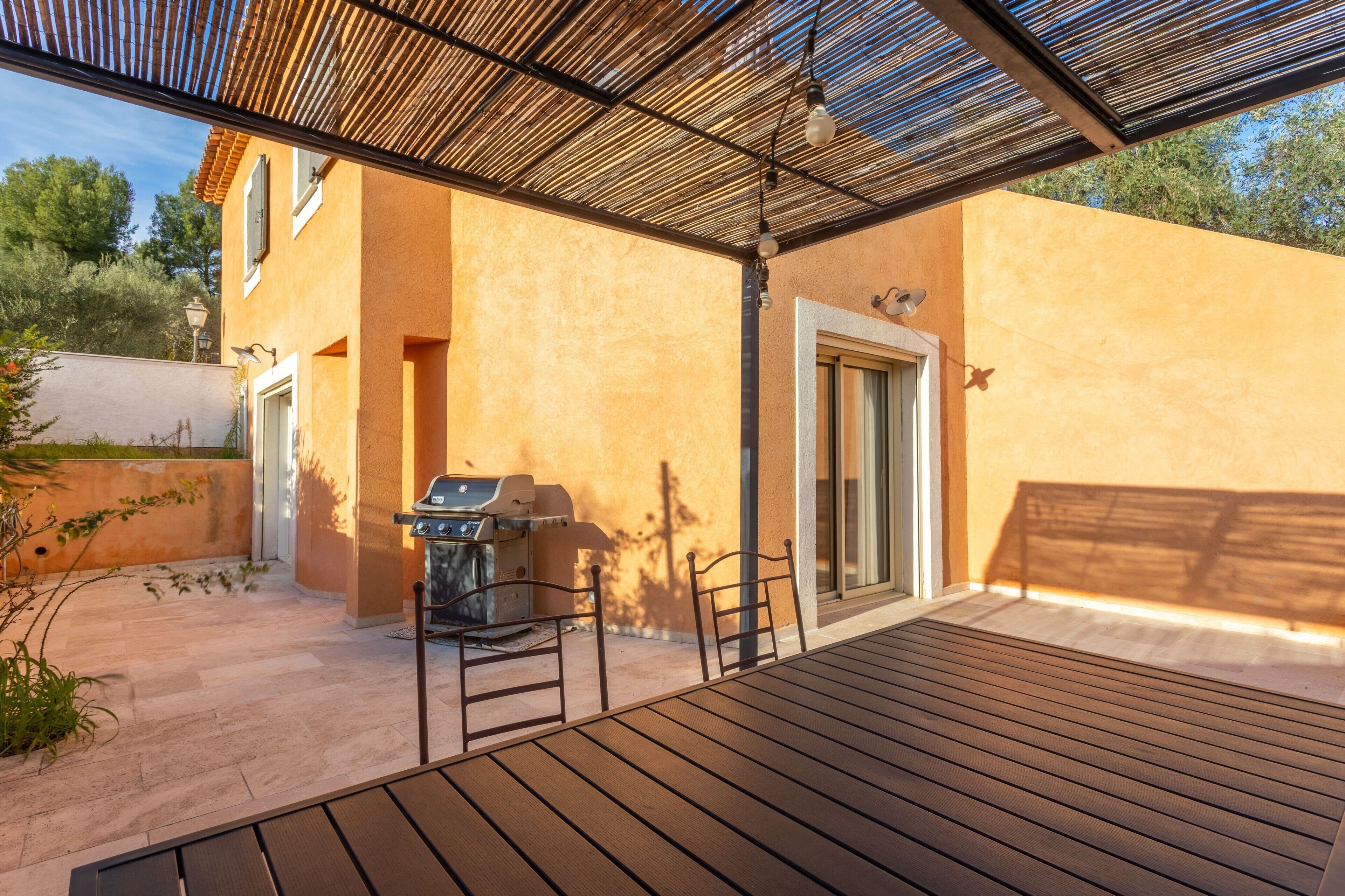 Een terras buiten met een barbecue, stoelen en een pergola.