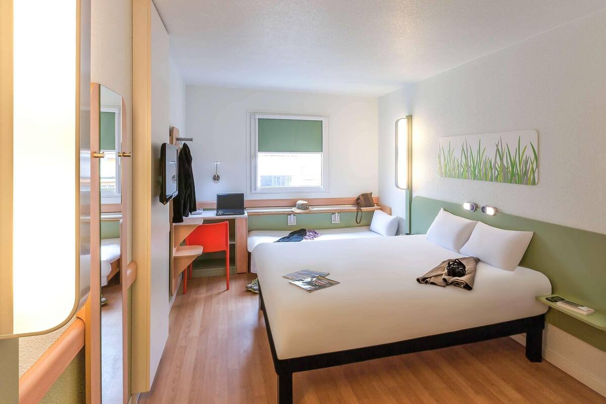 A hotel room with a bed, a desk with a laptop, a chair, a TV, and a window.