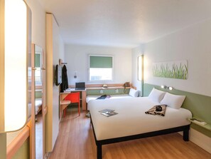 A hotel room with a bed, a desk with a laptop, a chair, a TV, and a window. - Ibis Budget Valencia Aeropuerto (Manises)
