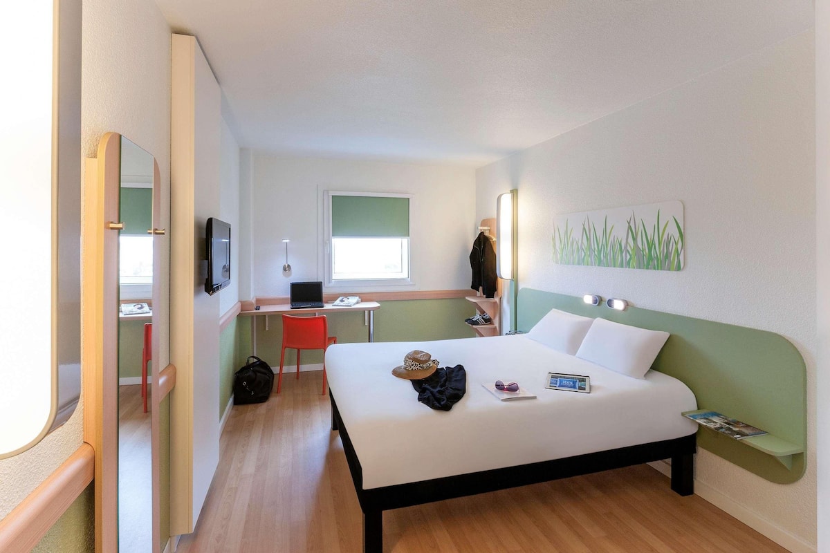A hotel room with a bed, a desk with a laptop, a chair, a TV, and a window.