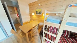 A compact room with a bunk bed, a wooden table with two chairs, a small kitchenette, and a single wall-mounted light. - Apartment in Plagne With Pool Access (La Plagne-Tarentaise)