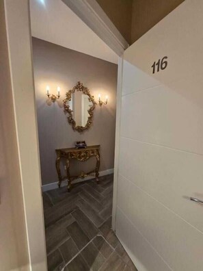 Un couloir avec un miroir, des appliques murales et une petite table. La pièce a un sol carrelé et une porte portant le numéro 116.