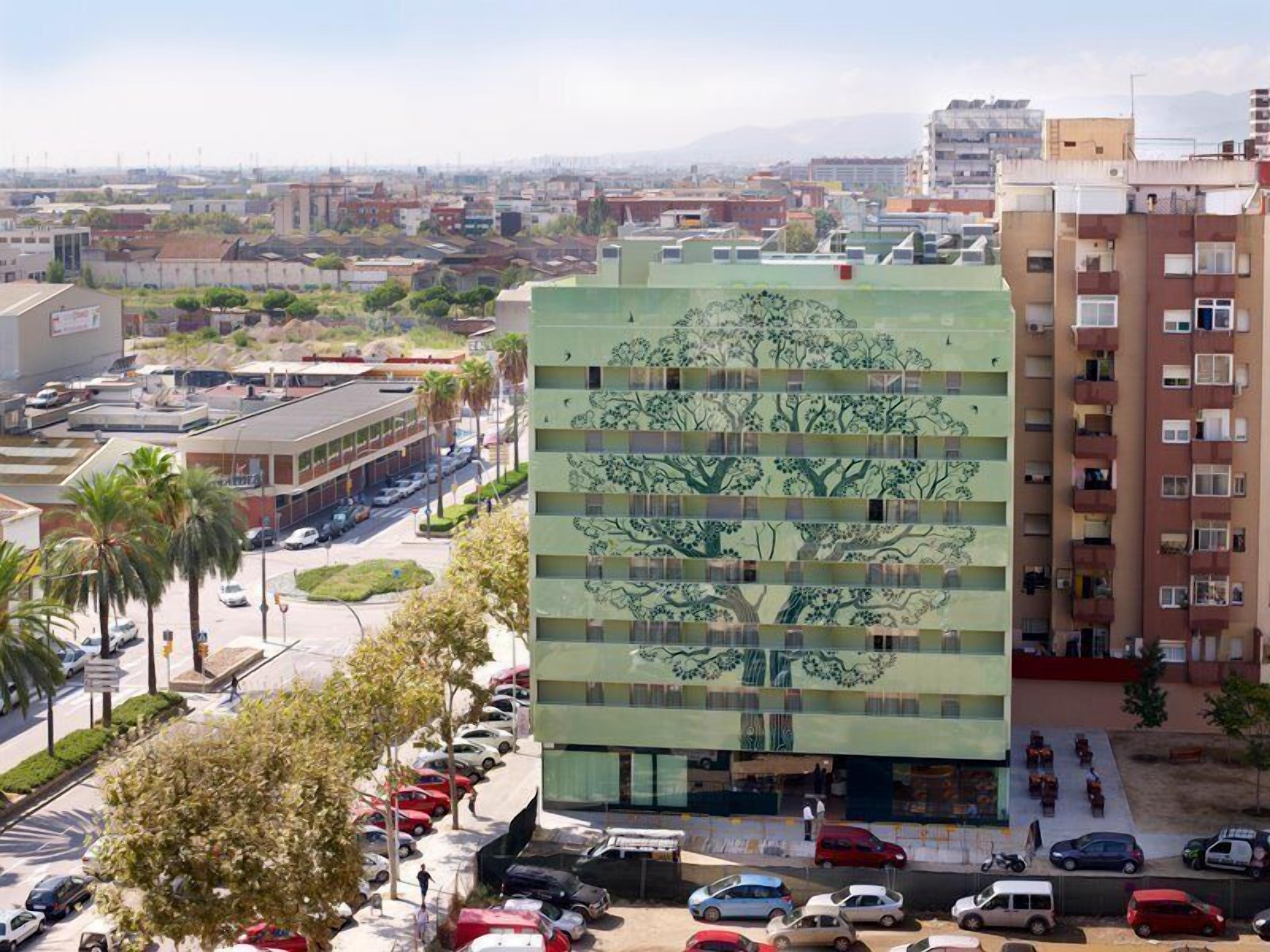 Un edificio moderno con un mural único de un árbol en su fachada, rodeado de un estacionamiento y otros edificios urbanos.