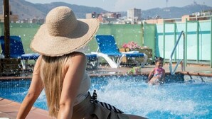 A poolside area with a person sitting on the edge, a towel, and a view of the pool and distant buildings. - Aura Park Aparthotel (L'Hospitalet de Llobregat)