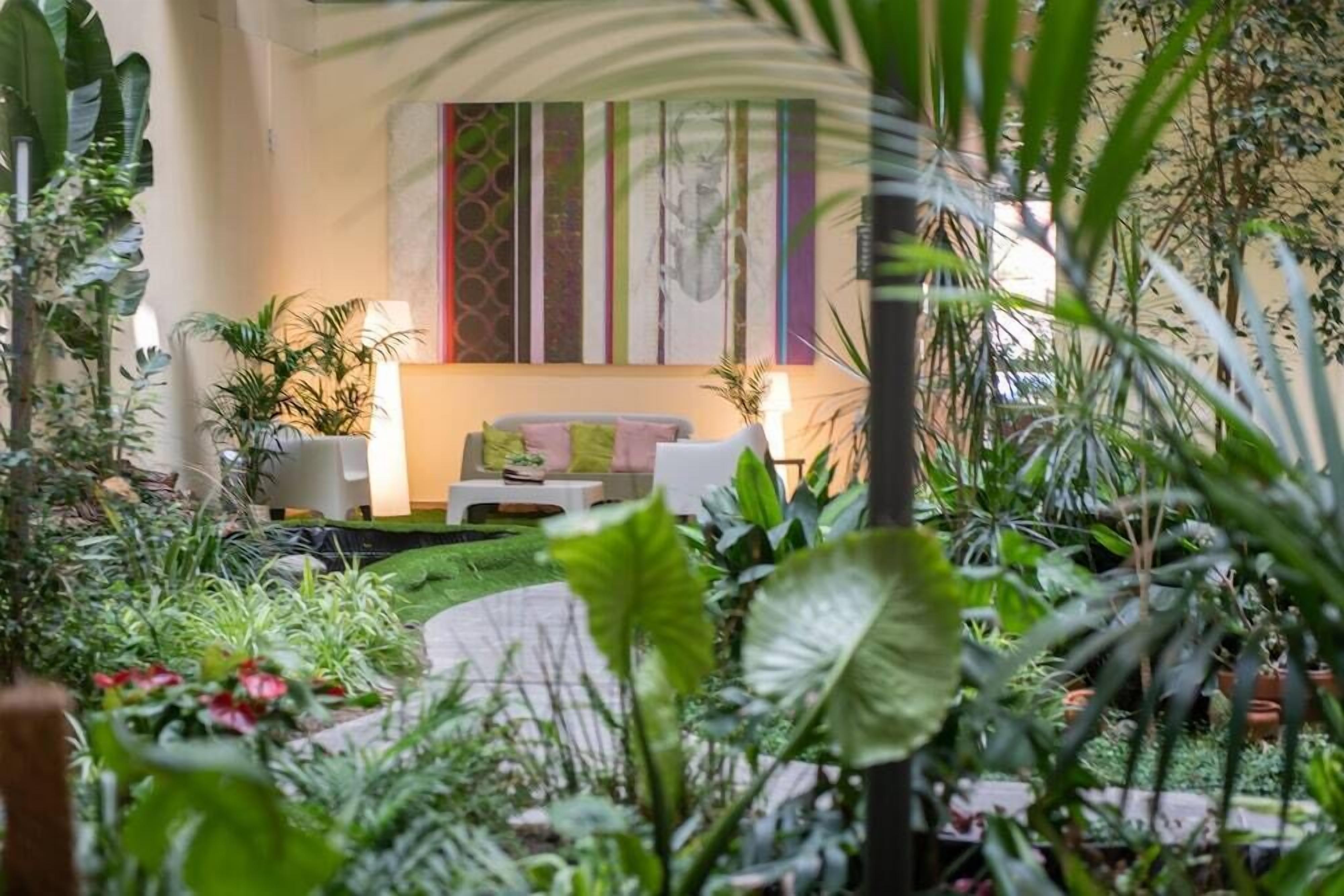 Un jardín interior acogedor con un sofá y sillones, rodeado de plantas exuberantes y un cuadro abstracto colorido en la pared.