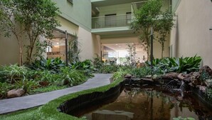 A courtyard with a pond, surrounded by lush greenery and a pathway. - Aura Park Aparthotel (L'Hospitalet de Llobregat)