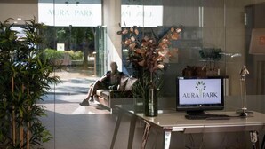 A reception desk with a computer displaying "AURA PARK" logo, a vase with flowers, and a person sitting outside. - Aura Park Aparthotel (L'Hospitalet de Llobregat)