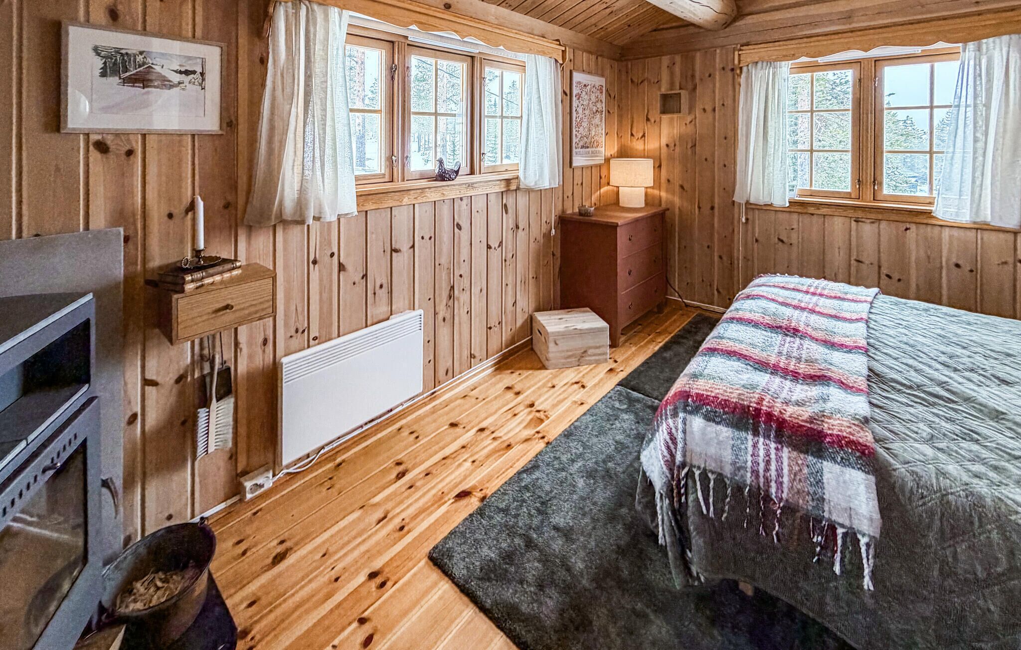 Ett mysigt rum i en stuga med träväggar, en säng med ett rutigt täcke, ett litet bord med en lampa och en öppen spis.