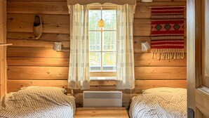 En hytte med to senge, et vindue med gardiner, en varmeovn og en lampe på væggen.
