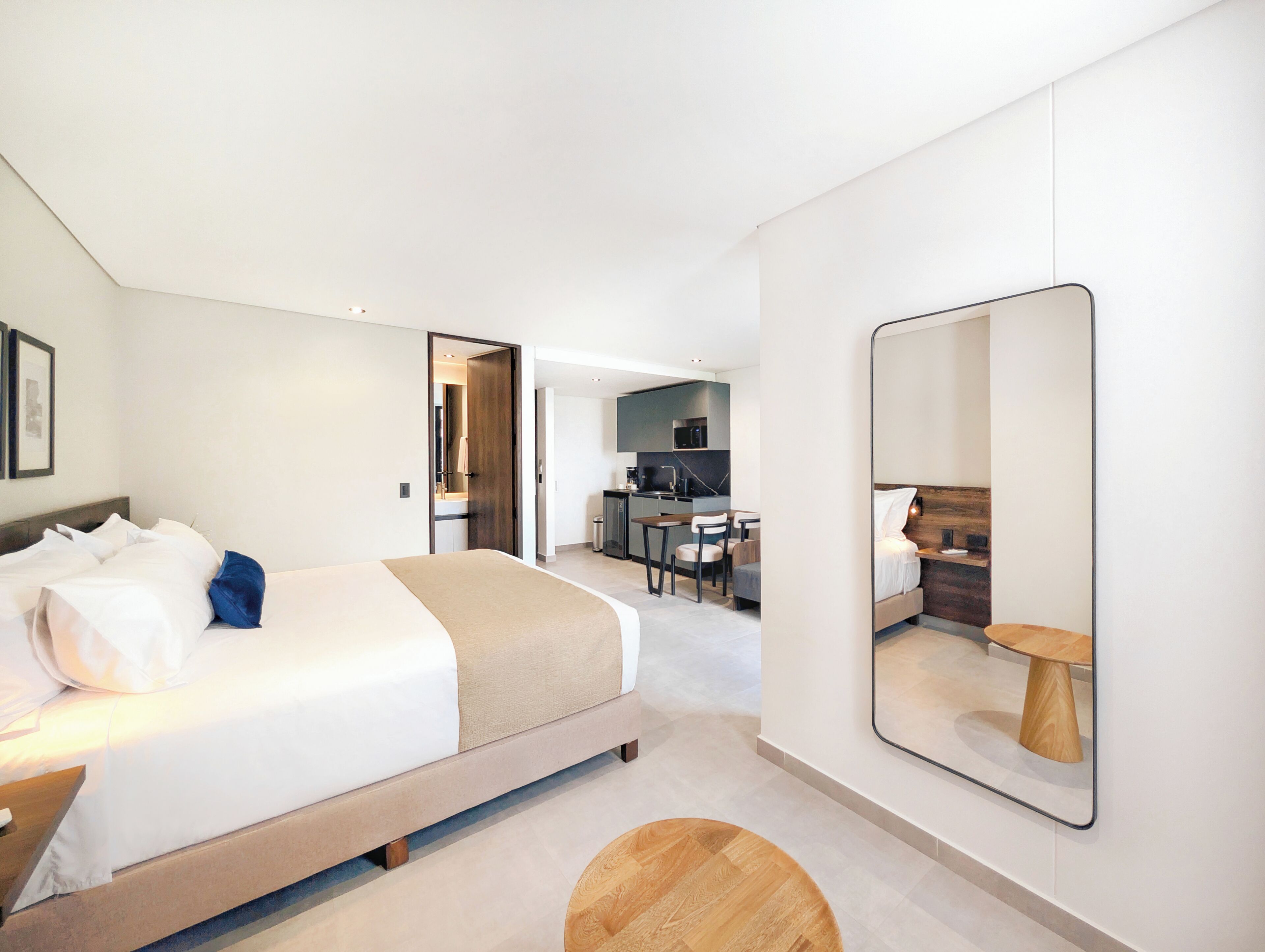 A modern hotel room with a bed, a desk, and a kitchenette in the background.
