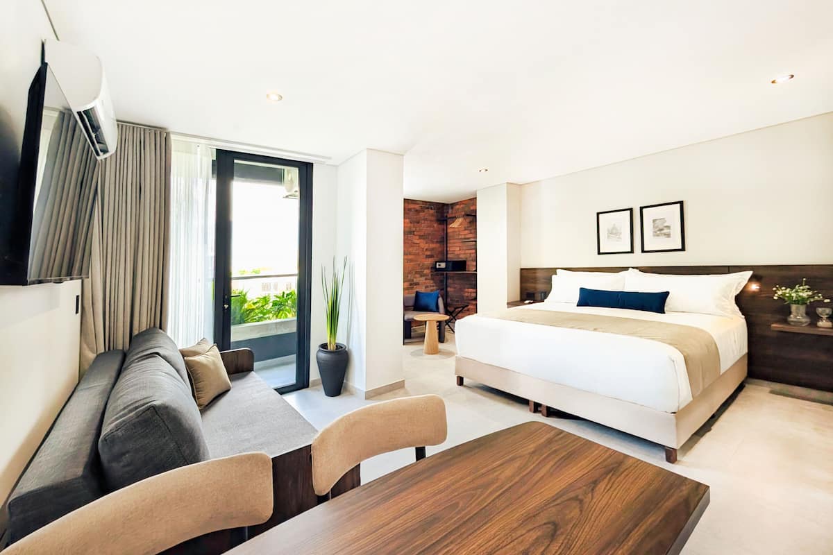 Ein modernes Hotelzimmer mit einem Bett, einer Sitzecke mit Sofa und Sesseln, einem Esstisch und einem Balkon mit Vorhängen.