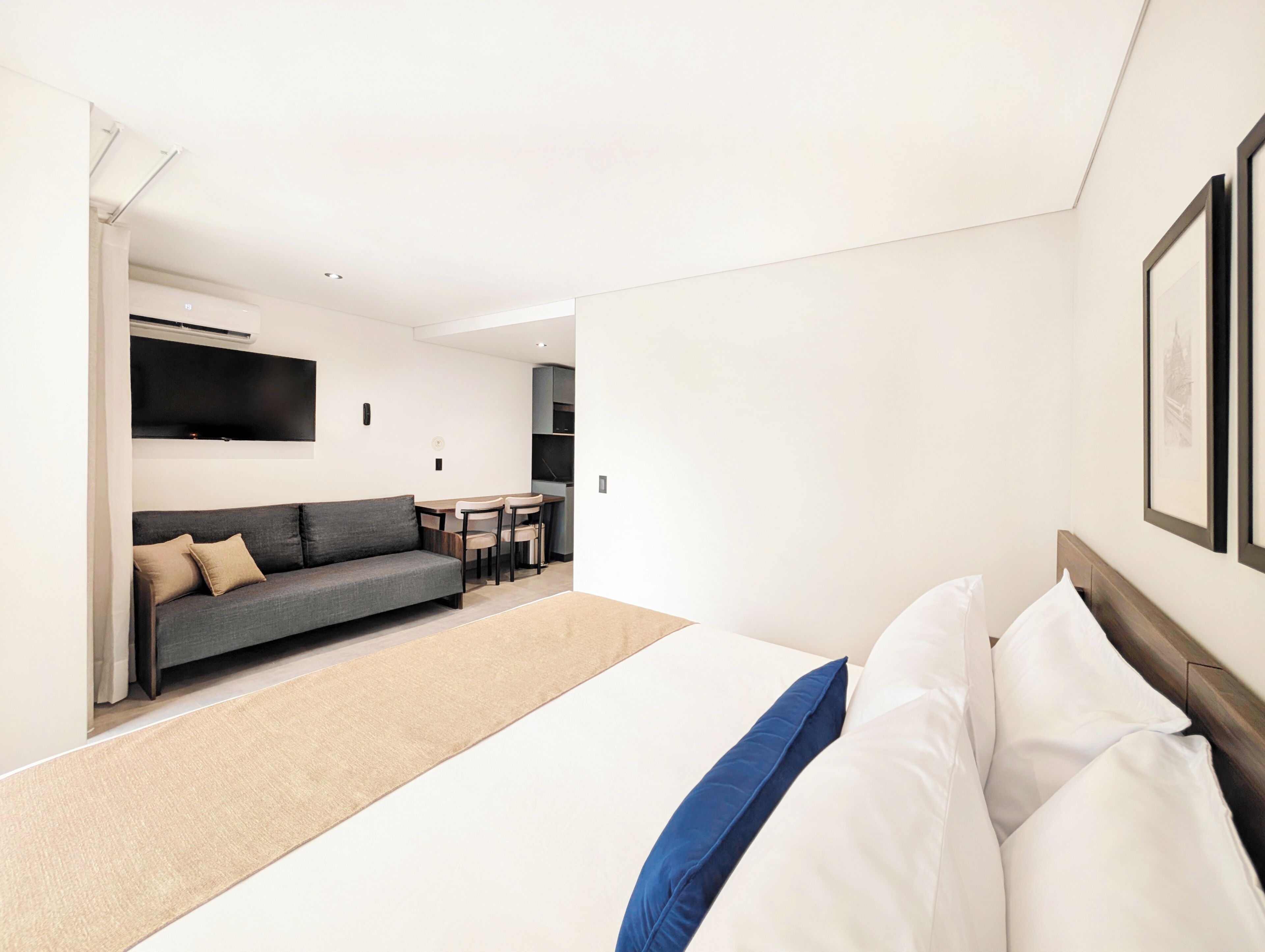 A modern hotel room with a large bed, a sofa, a dining table, and a flat-screen TV mounted on the wall.