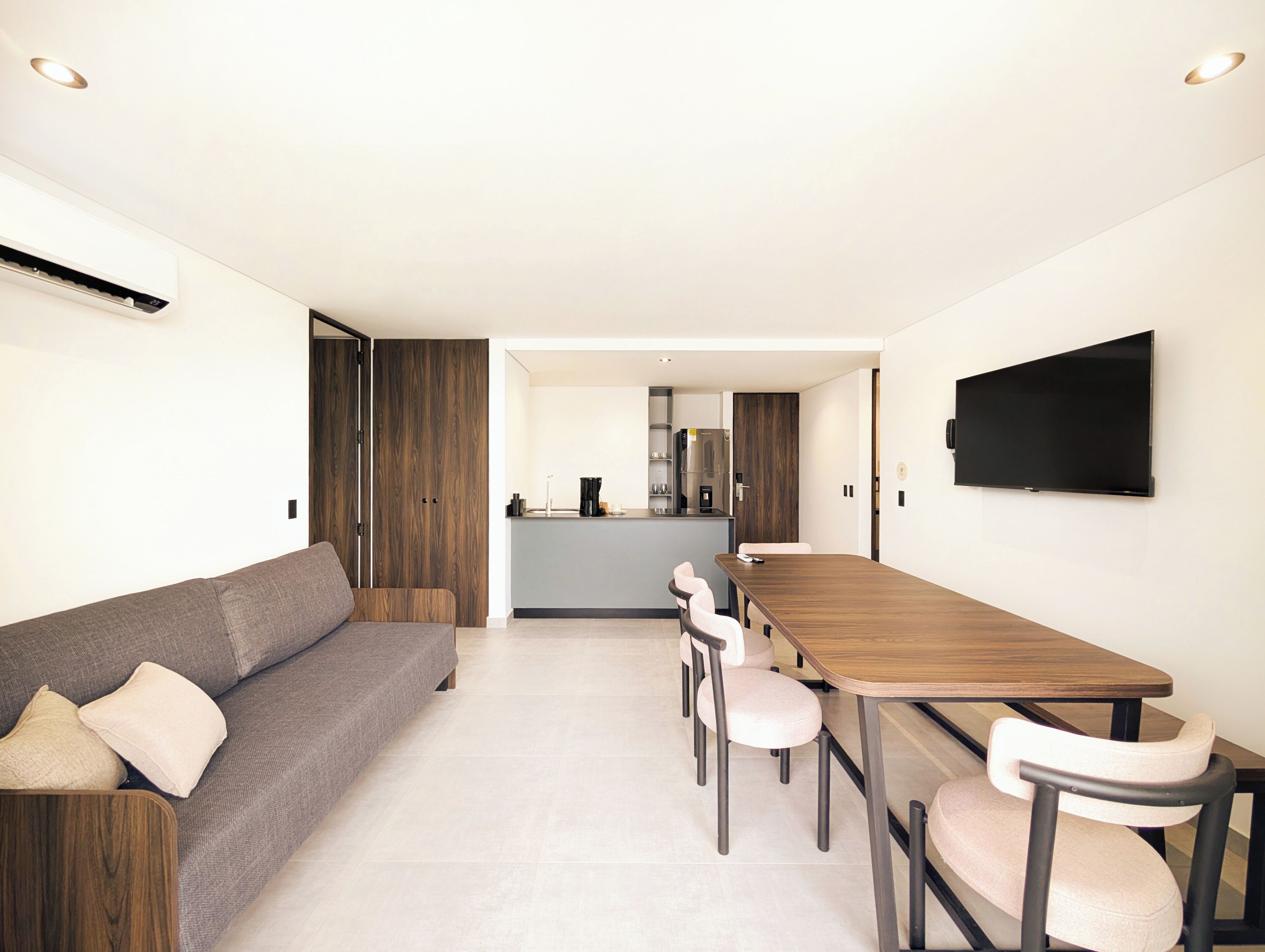 A modern living room with a sofa, dining table, and chairs, a kitchen in the background, and a TV mounted on the wall.