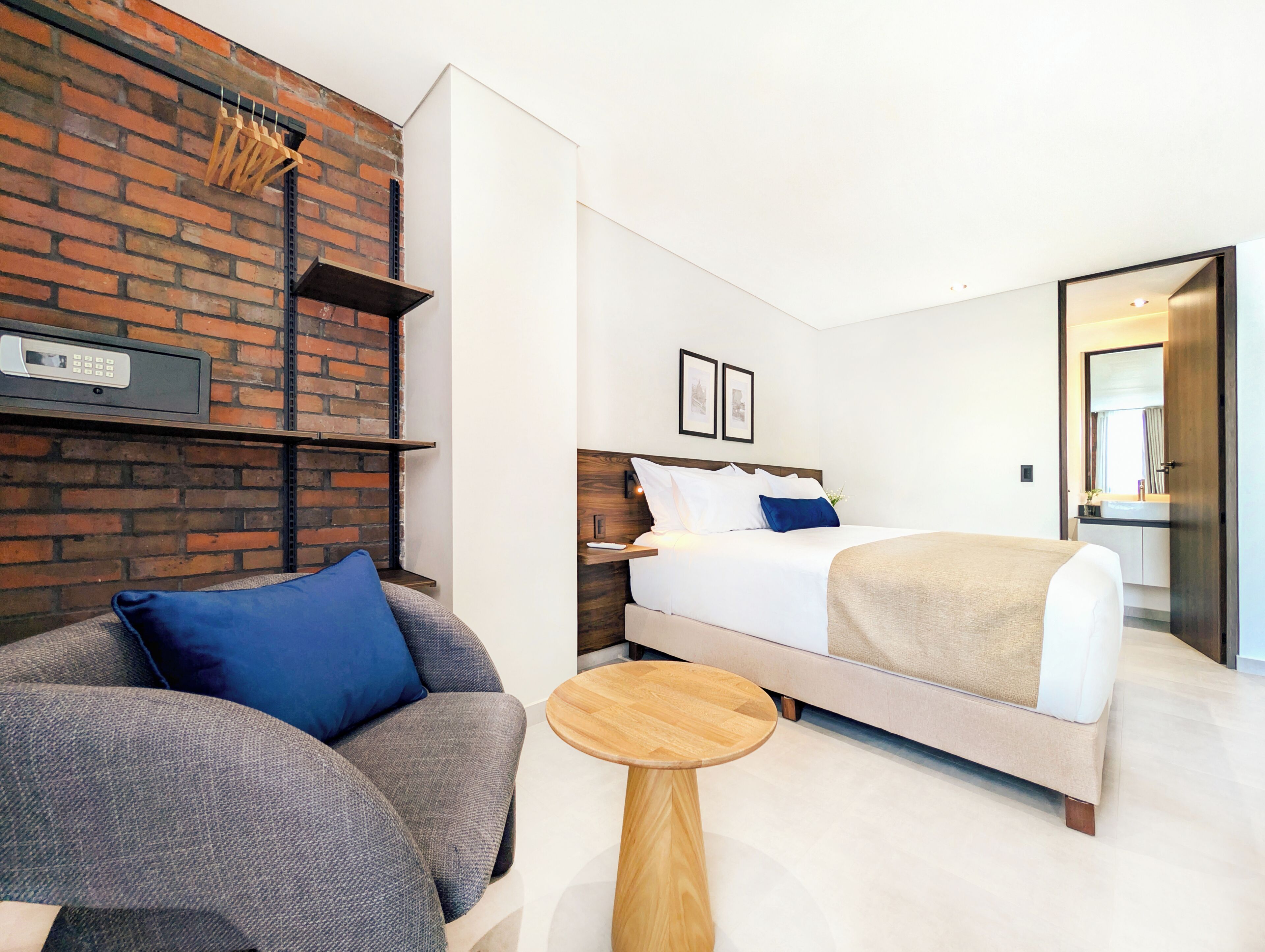 Una camera d'albergo moderna con una parete decorata in mattoni, un letto con biancheria da letto bianca, un divano grigio con cuscini blu e un piccolo tavolino in legno.