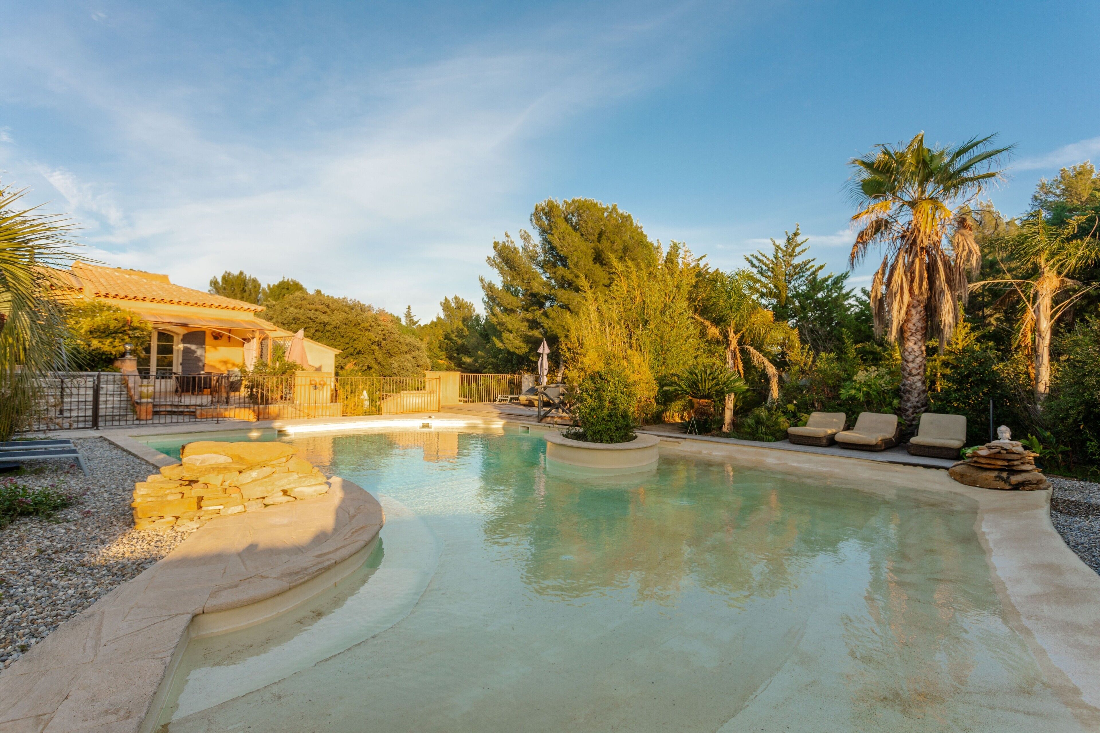 Une piscine à bord incurvé, entourée de palmiers et bordée d’un bâtiment dont le toit est recouvert de tuiles.