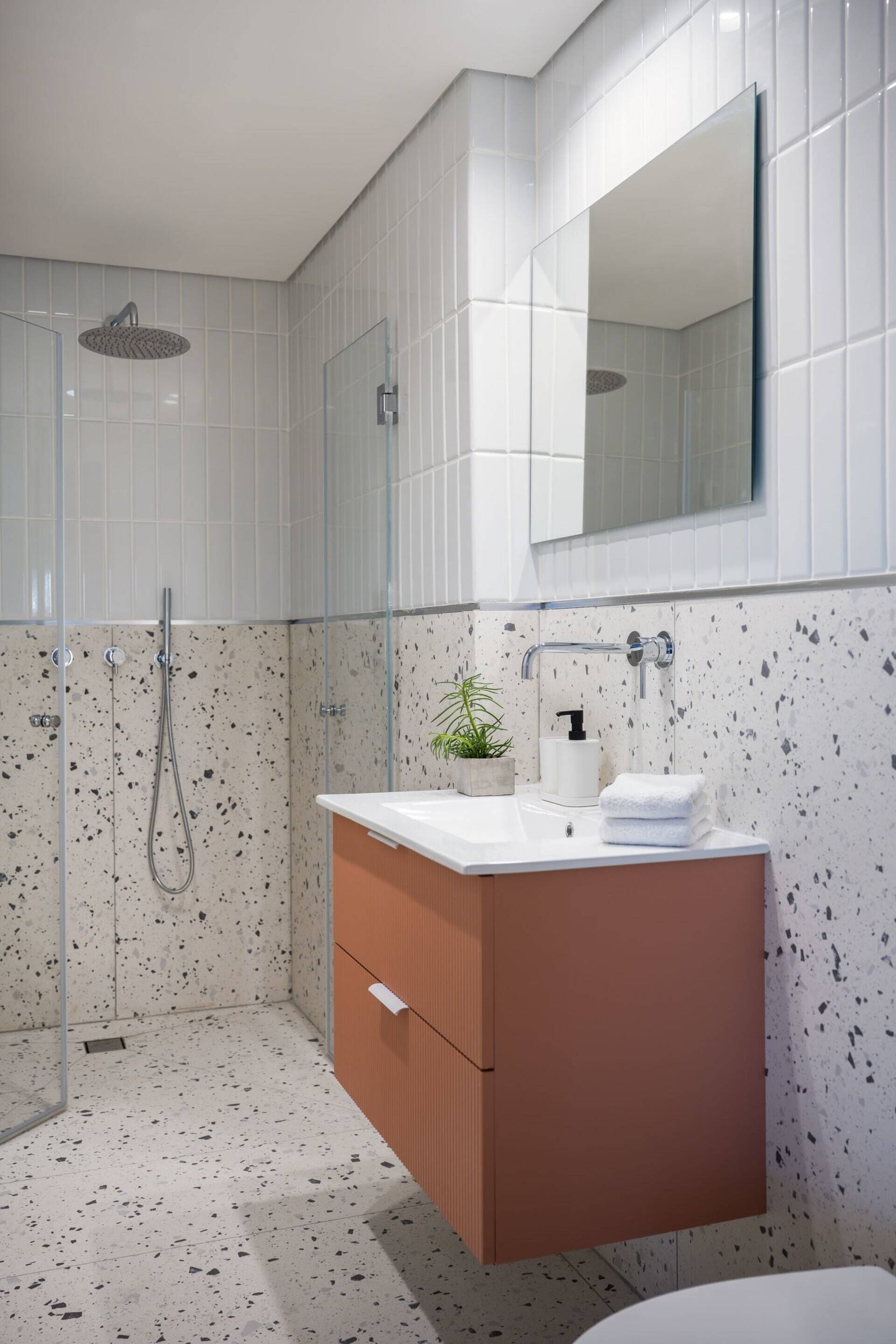 Ein modernes Badezimmer mit einer gläsernen Dusche, einem weißen Waschtisch mit Spiegel und Wandfliesen mit Farbflecken.