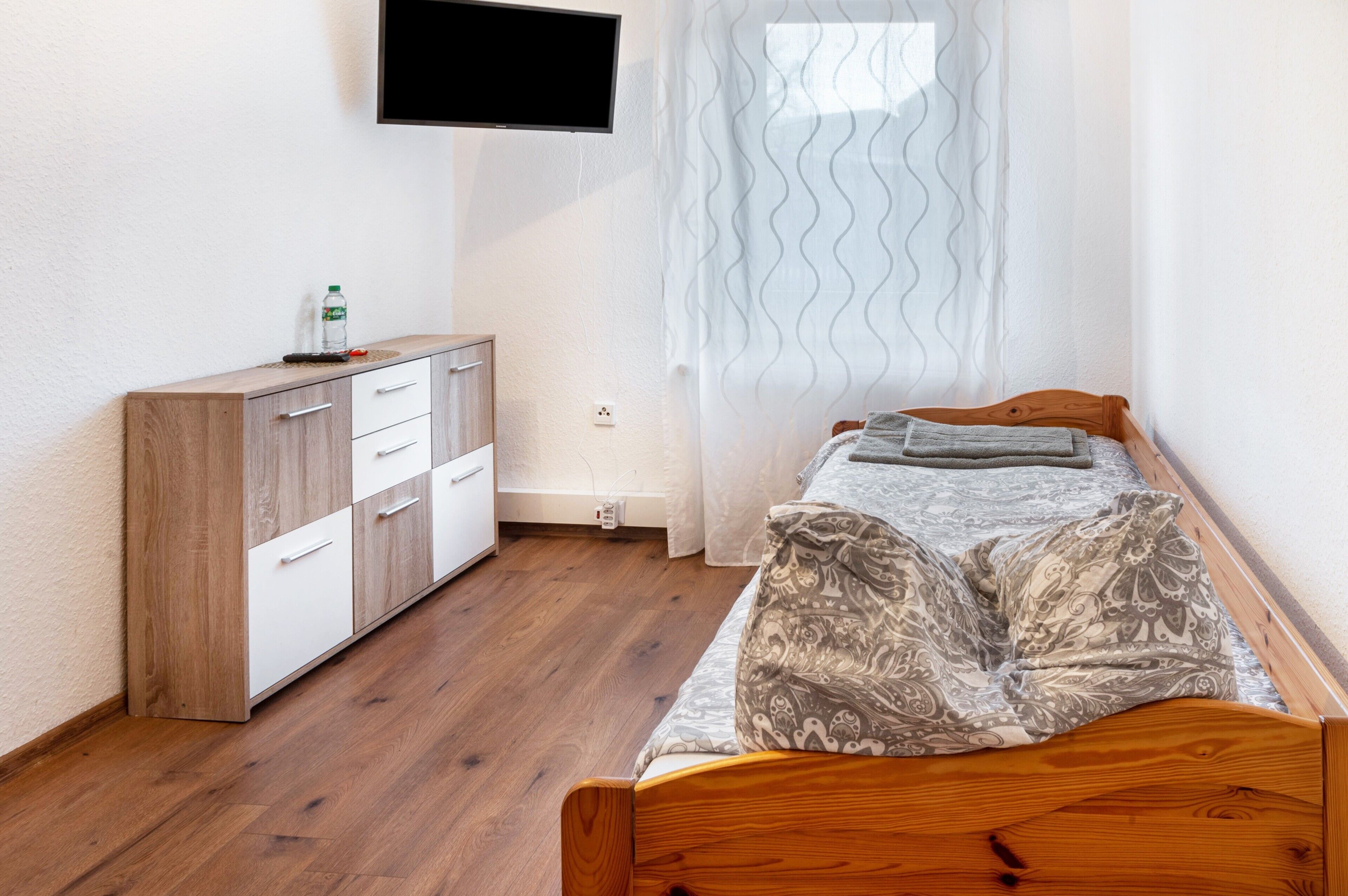 A bedroom with a wooden bed, a white dresser, a flat-screen TV mounted on the wall, and a window with sheer curtains.