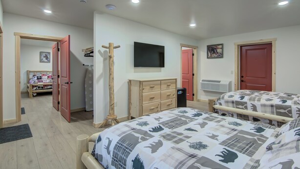 A bedroom with two beds, a wooden dresser, a TV mounted on the wall, and a red door. - Lundy's Black Bear - Blair Oaks (Drummond)