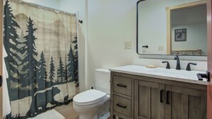 A bathroom with a white toilet, a wooden vanity with drawers, a large mirror, and a shower curtain with a forest design. - Lundy's Black Bear - Blair Oaks (Drummond)