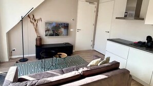 A modern living room with a sofa, a coffee table, a TV, and a kitchen area with cabinets and a countertop. - Lodge Located on the Church Square (Zoutelande)