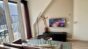 A living room with a sofa, a flat-screen TV, a small side table, and a floor lamp. - Lodge Located on the Church Square (Zoutelande)