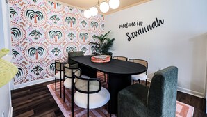 Ein Essbereich mit einem runden Tisch, Stühlen, einer Wand mit dem Schriftzug 'meet me in Savannah', strukturiertem Wandpapier und einer Pflanze.