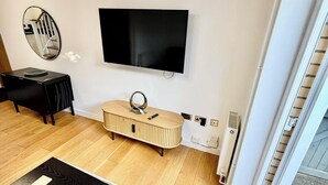 A modern living room with a flat-screen TV mounted on the wall, a wooden console table, a black sofa, and a patterned rug. - 2 Bed House - Sleeps 4 - Parking - Wifi (Cambridge)