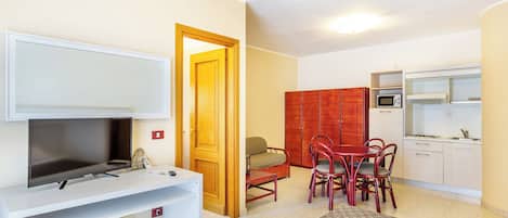 A compact living space with a TV on a white stand, a dining area with red chairs, a small kitchen with a microwave, and a red cabinet.