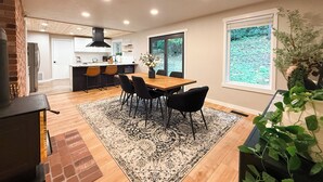 Une cuisine moderne dotée d’un îlot central, d’un coin repas avec une table en bois et des chaises noires, et d’une grande fenêtre offrant une vue sur la verdure.