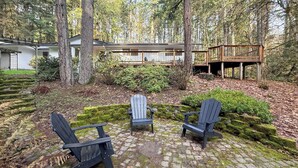 Une terrasse en bois, agrémentée de fauteuils Adirondack, entourée d’une zone boisée, avec une maison en arrière-plan.