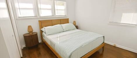 Un dormitorio con cama de madera, ropa de cama blanca, una mesita de noche con lámpara y una ventana con persianas.