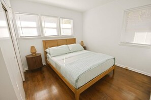 A bedroom with a wooden bed, white bedding, a nightstand with a lamp, and a window with blinds. - Walkable to Notre Dame! Cozy 2 bedroom home(426) (South Bend)