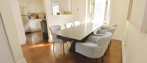 A dining area with a long table and six chairs, a kitchen in the background, and a window providing natural light.
