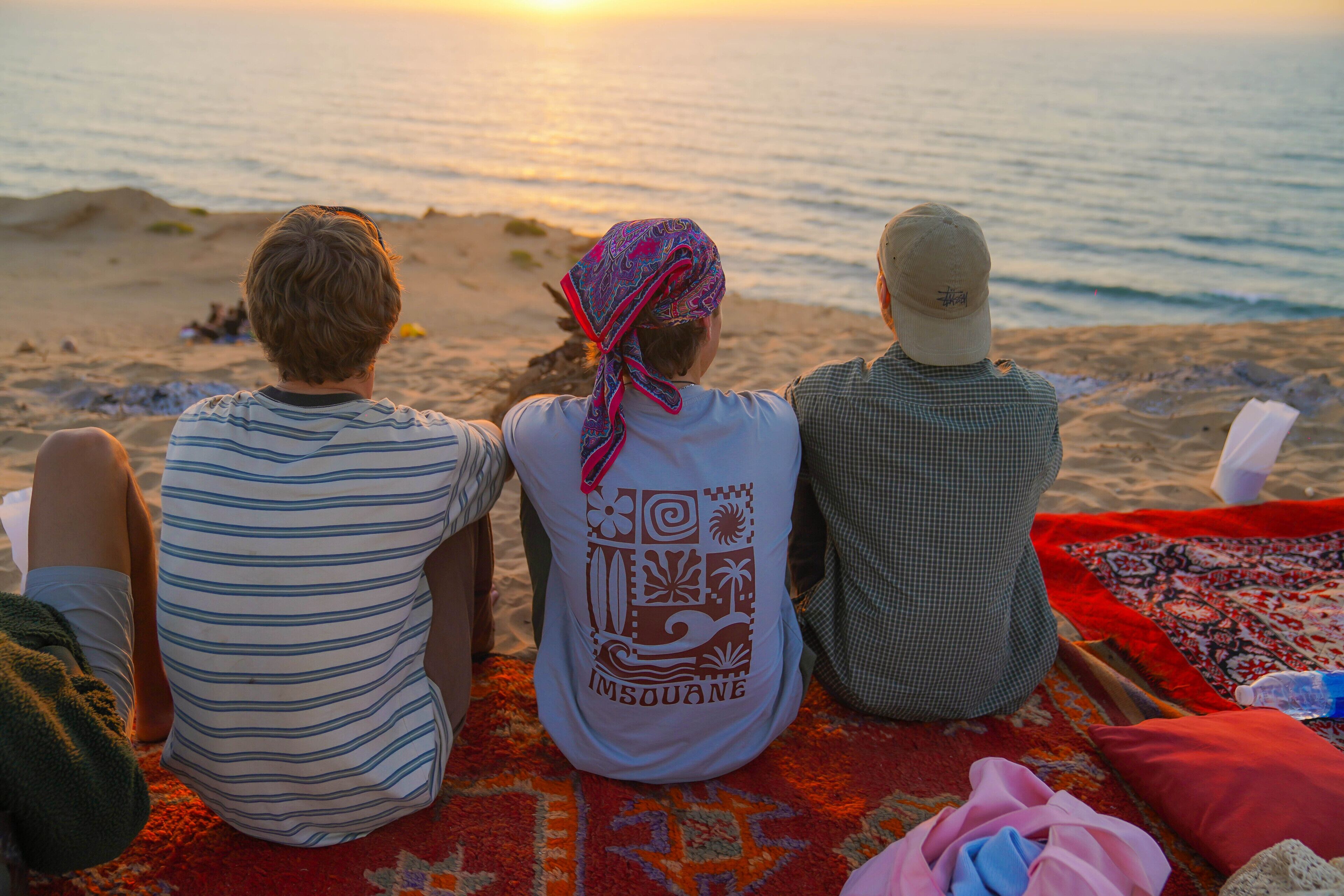 Drei Personen sitzen auf einer Decke am Strand, im Hintergrund ein Sonnenuntergang.