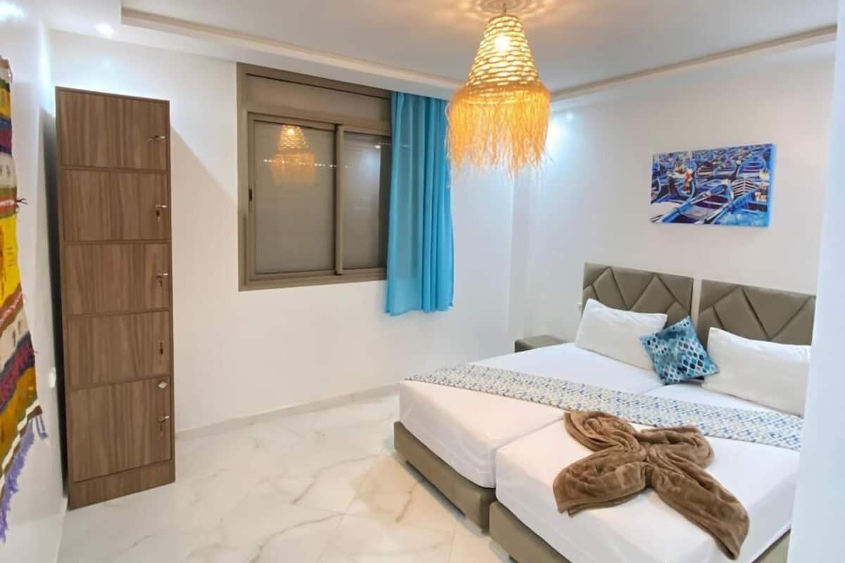 A bedroom with a bed, a wardrobe, a window with curtains, a painting on the wall, and a hanging light fixture.