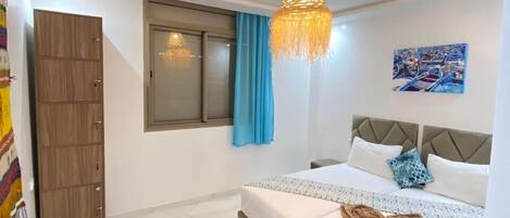 Un dormitorio con cama, armario, ventana con cortinas, un cuadro en la pared y una lámpara colgante.