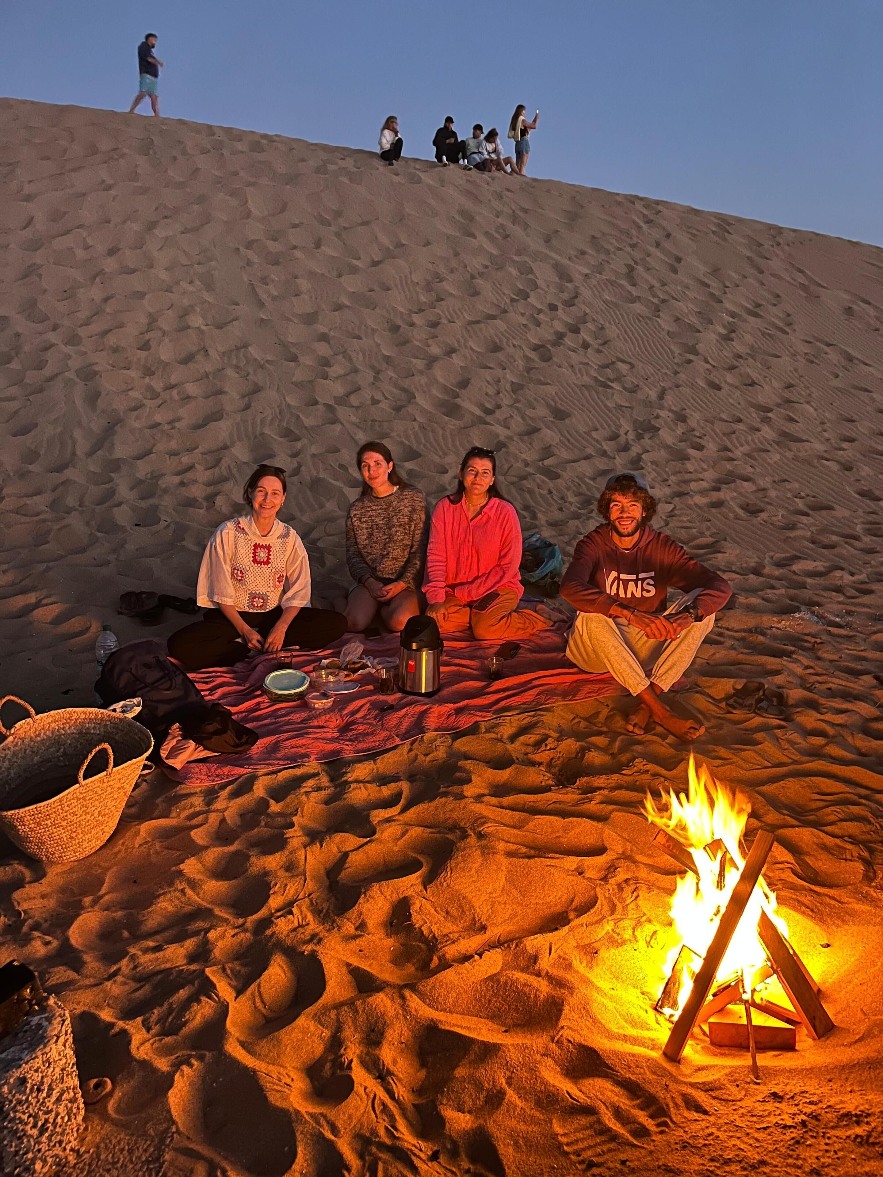 A group of people sitting on a blanket in a sandy area with a campfire.
