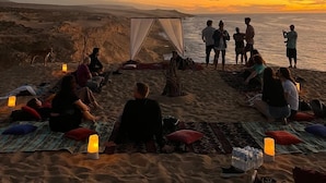 Eine Gruppe von Menschen sitzt auf Decken an einem sandigen Strand bei Sonnenuntergang, mit einem Zelt und einigen Personen, die am Wasser stehen.