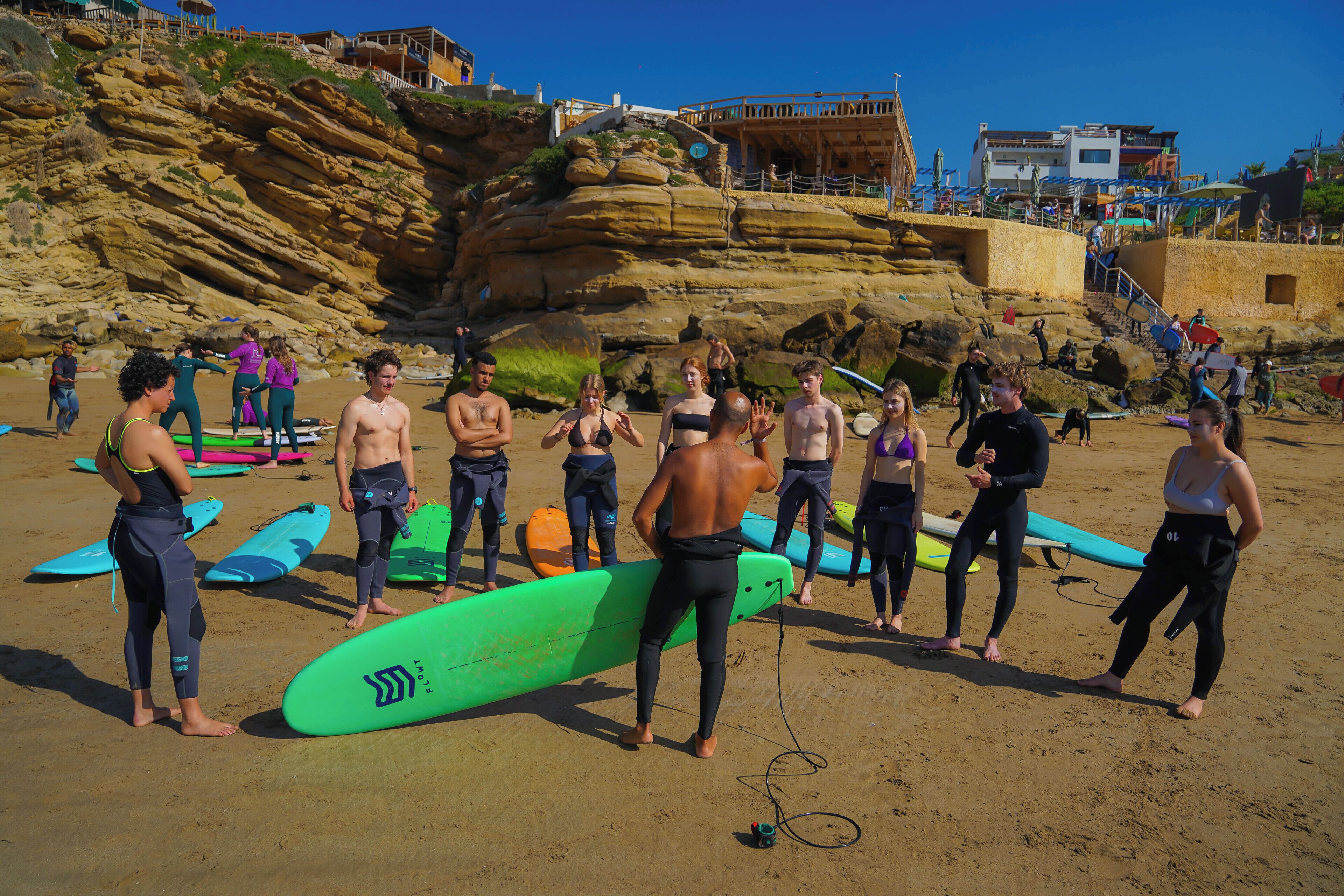 Ein Strand mit Menschen in Neoprenanzügen und Surfbrettern, felsigen Klippen und Gebäuden im Hintergrund.