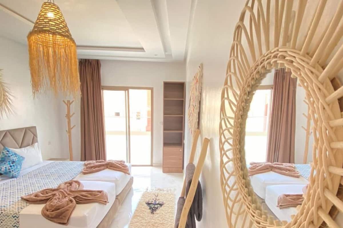 A modern bedroom with a large bed, a hanging light fixture, a decorative mirror, and a view of the outside.