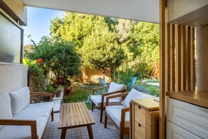 A covered patio with wooden furniture, a small table, and a view of a garden with trees and plants.