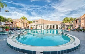 A swimming pool with a 5 ft depth marker, surrounded by lounge chairs and umbrellas, with buildings in the background. - Mickey's Hideaway Condo Near Disney (Kissimmee)
