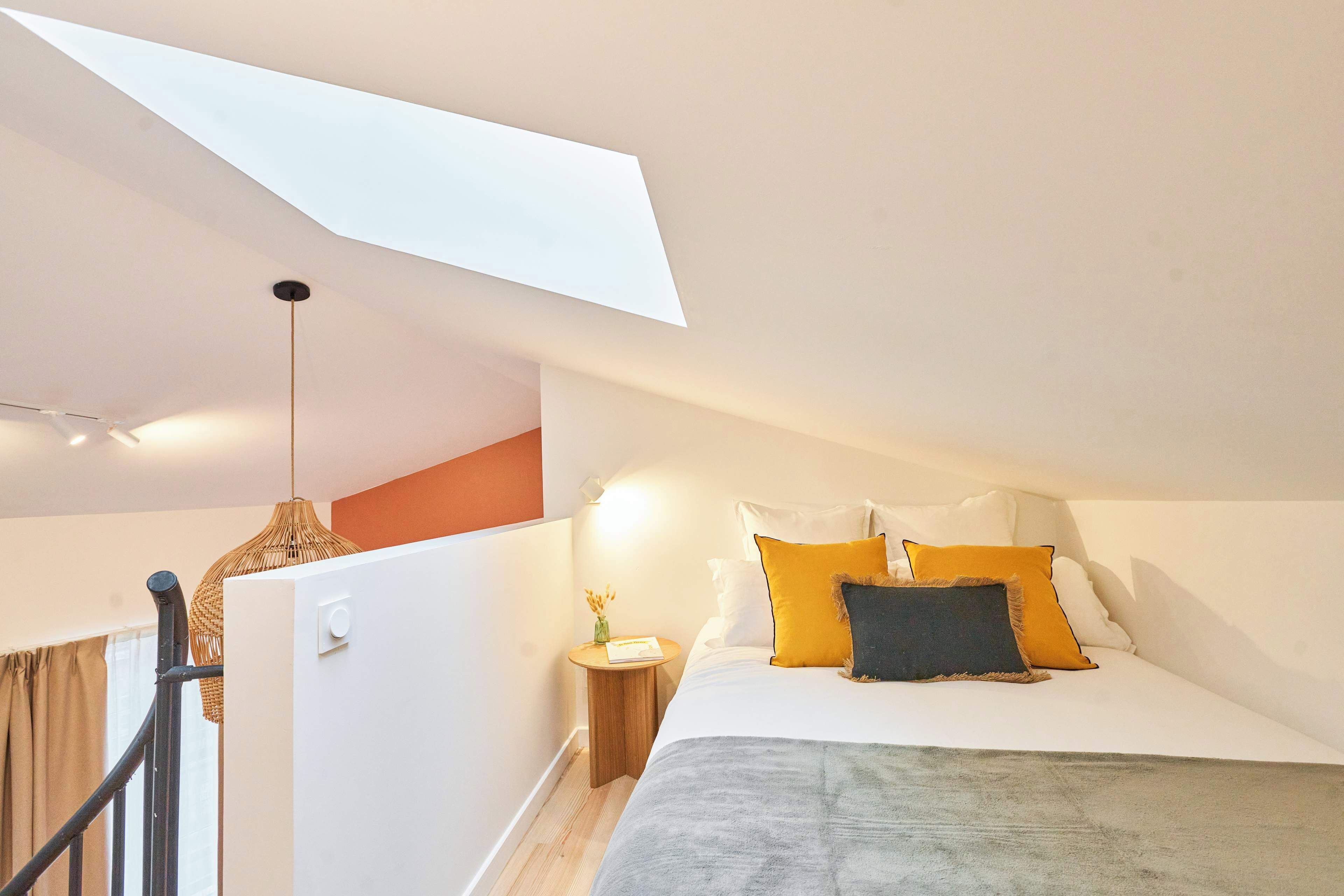 A bedroom with a skylight, a bed with multiple pillows, a bedside table with a lamp, and a staircase with a railing.