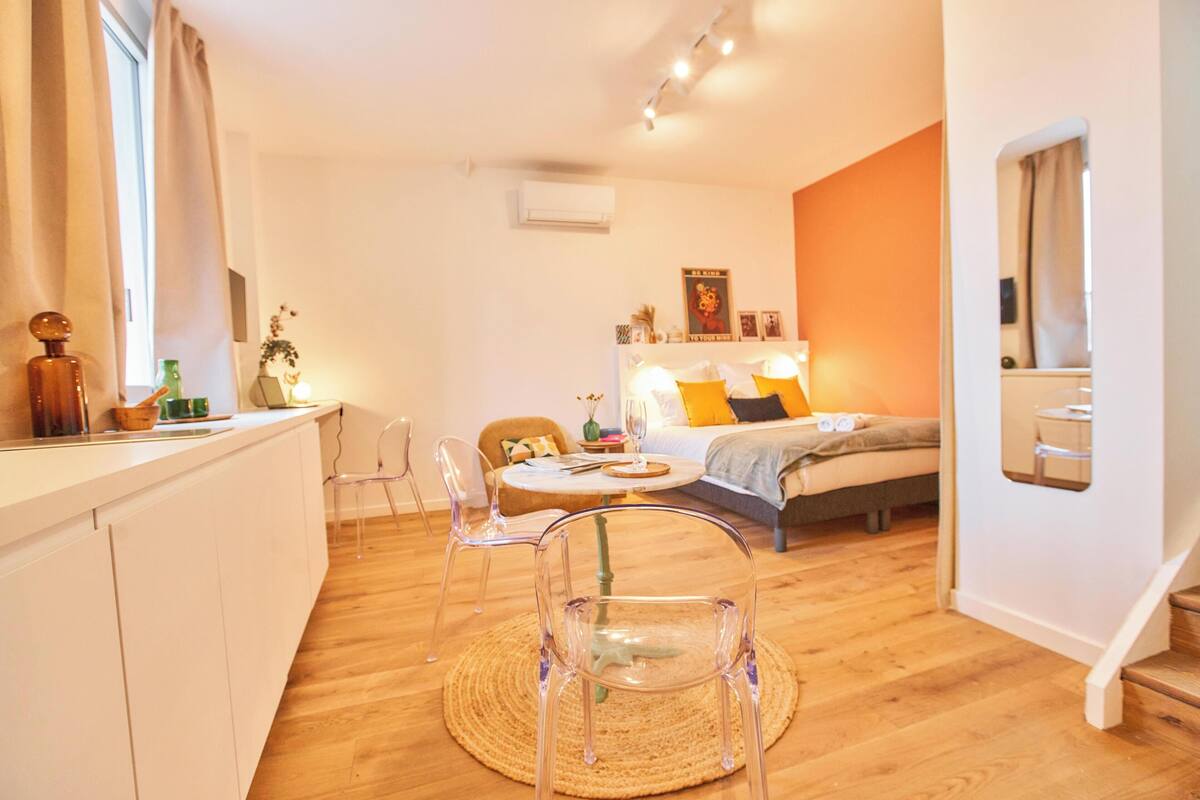 Ein modernes, helles Zimmer mit Holzboden, einem Esstisch mit Stühlen, einem Bett mit Kissen und einer kleinen Küchenzeile.