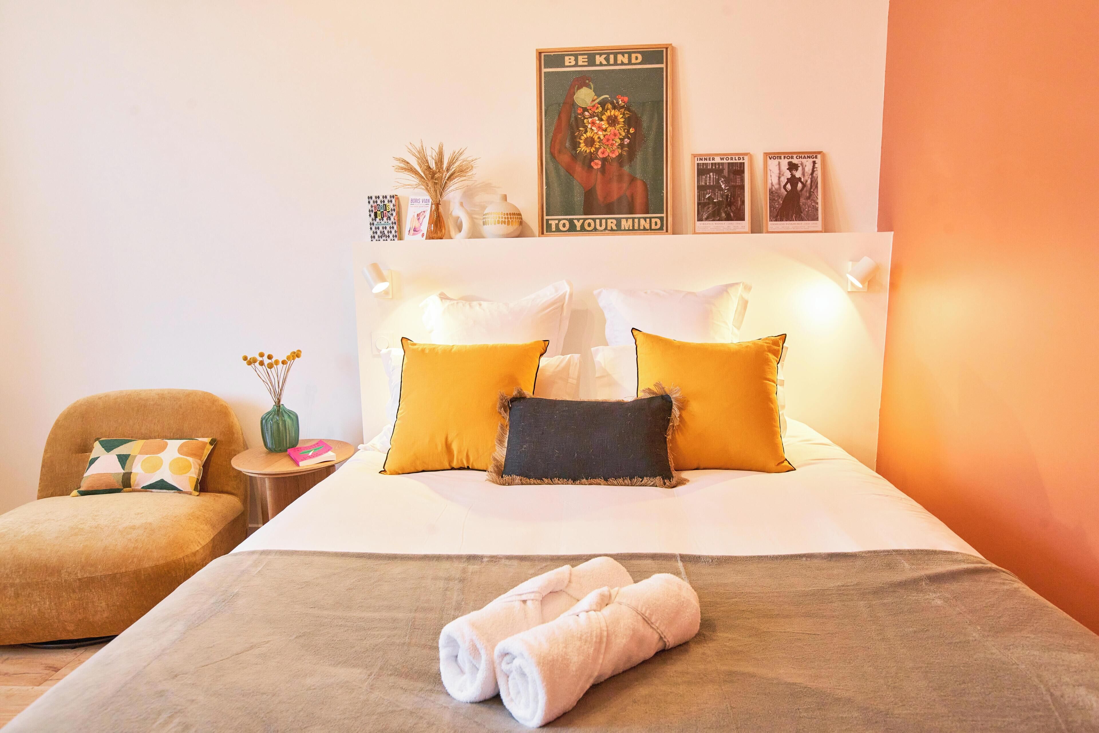 A neatly made bed with rolled towels, a headboard with multiple pillows, a small side table with a vase, and a framed inspirational poster on the wall.