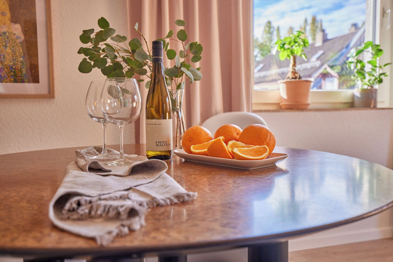 Une table ronde avec une bouteille de vin, deux verres, une assiette d’oranges et une serviette pliée.