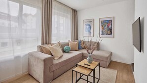 A modern living room with a beige sofa, a small side table, and a vase of branches. There are two framed artworks on the wall and a television mounted on the wall. - Bella - Kaiserslautern (Kaiserslautern)