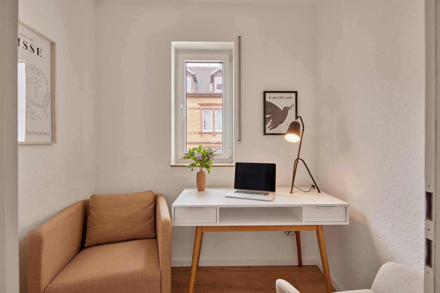 Una habitación pequeña y bien iluminada con un escritorio blanco, una laptop, una planta en maceta, una lámpara y un cuadro en la pared.